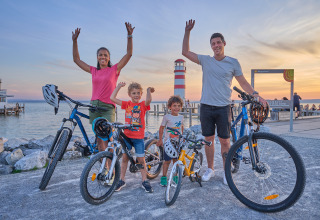 Famiglia che posa con le biciclette vicino all'acqua al tramonto accanto a un faro a Strandcamping Podersdorf am See, Austria.