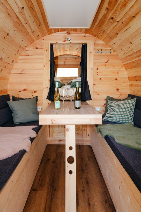 Gemütlicher Innenraum eines Weinfasses als Glamping-Unterkunft mit Tisch, Weinflaschen und Kissen.