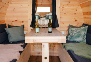 Gemütlicher Innenraum eines Weinfasses als Glamping-Unterkunft mit Tisch, Weinflaschen und Kissen.