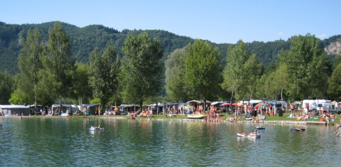 Campeggio sul lago con molte persone, tende e roulotte, circondato da alberi e colline verdi.
