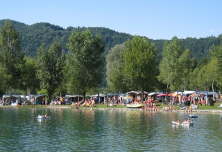 Camping aan een meer met veel mensen, tenten en caravans, omgeven door bomen en groene heuvels.