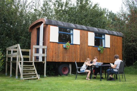Familie sitzt draußen bei einem Holz-Pipowagen auf Familiecamping Hendriks Wijkje in Drenthe, Niederlande.