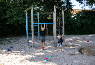 Zwei Kinder spielen auf einem Spielplatz mit Klettergerüst im Sand bei Familiecamping Hendriks Wijkje - Pipowagens Drenthe.