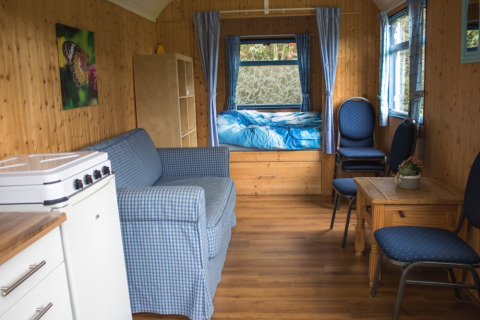 Innenansicht einer gemütlichen Pipowagen mit Holzwänden, Bett, Stühlen, Sofa und kleinem Küchenbereich.