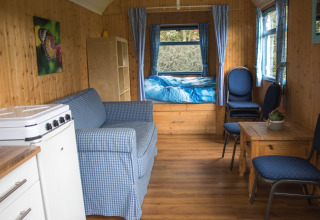 Interieur van een gezellige pipowagen met houten wanden, bed, stoelen, bank en een kleine keukenhoek.
