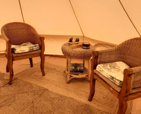 Alojamiento glamping acogedor con dos sillas de mimbre, mesa pequeña, libros y mantas dentro de una tienda.