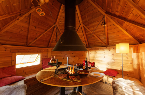 Indvendigt af en hyggelig træyurt med centralt ildsted, spisebord og varme omgivelser i glampingstil.