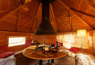 Interno di una yurta in legno accogliente con fuoco centrale, tavolo da pranzo e atmosfera glamping calda.