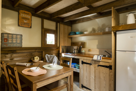 Gezellige glampingkeuken met houten meubels, eettafel, open planken, koelkast en een raam met uitzicht.