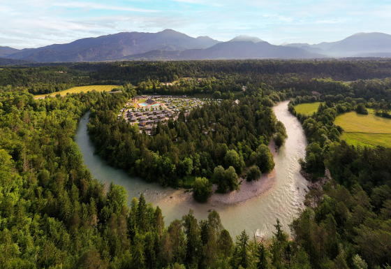 Vue aérienne de River Camping Bled - Lodges Slovenië avec une rivière, forêt et montagnes à l’arrière-plan.