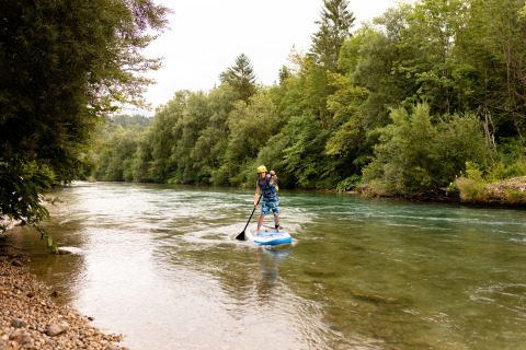 Person paddelt auf einem Stand-Up-Board auf dem Fluss bei River Camping Bled - Lodges Slovenië, umgeben von Wald.