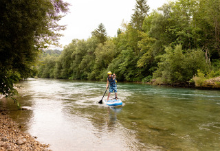 Person paddleboarding on a river at River Camping Bled - Lodges Slovenië, surrounded by lush green forest.