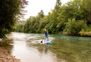 Persoon op een paddleboard op de rivier bij River Camping Bled - Lodges Slovenië, omgeven door natuur.