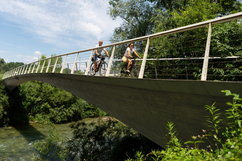 Zwei Personen radeln über eine Brücke inmitten grüner Natur bei River Camping Bled - Lodges Slovenië.