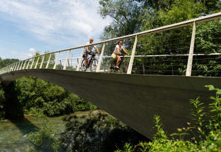 Two people biking across a bridge surrounded by lush greenery at River Camping Bled - Lodges Slovenië.