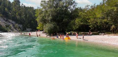 Folk nyder en solrig dag ved floden på River Camping Bled - Lodges Slovenië omgivet af skov.