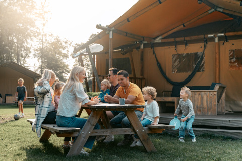 Famille réunie à une table en bois devant une tente de glamping à Vakantiepark de Italiaanse Meren, Gelderland.