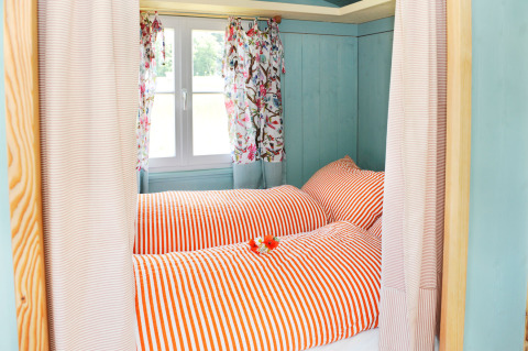 Habitación de glamping con ropa de cama a rayas rojas y cortinas florales en Hofgut Hopfenburg, Baden-Württemberg.
