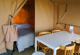 Vista interior de una tienda glamping con zona de dormitorio, mesa de comedor y sillas sobre suelo de madera en Ameland.