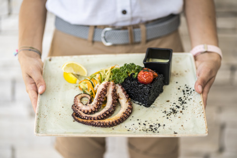 Una persona sirve pulpo a la parrilla, arroz negro, verduras, limón y salsa, presentado en un plato elegante.