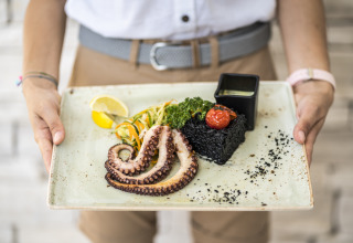 Une personne sert du poulpe grillé, du riz noir, des légumes, du citron et une sauce joliment dressés.