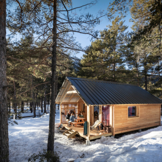Glampingverblijf Huttopia Winter Chalets – La Clarée, houten hut in besneeuwd bos in de Franse Alpen.