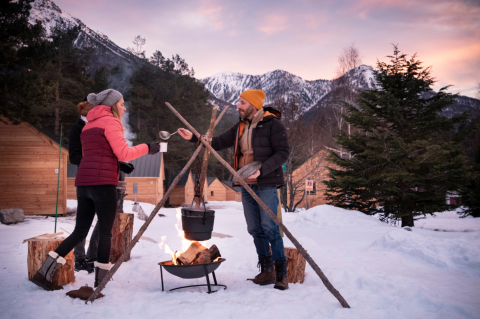 Winter glamping at Huttopia Winter Chalets – La Clarée in the French Alps, people around a snowy campfire.