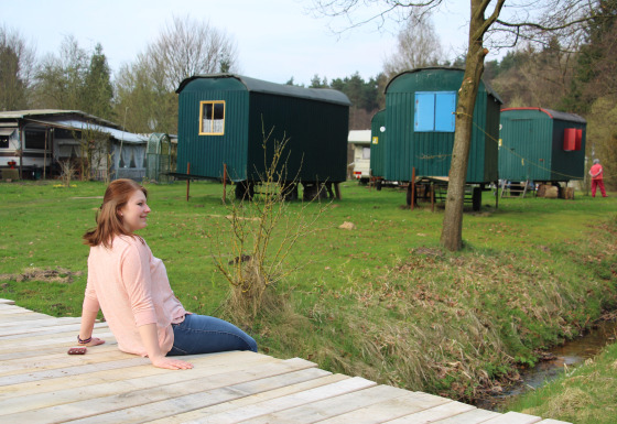 Femme assise sur plateforme en bois au Camping Böhmeschlucht avec pipowagens verts en arrière-plan en Basse-Saxe.