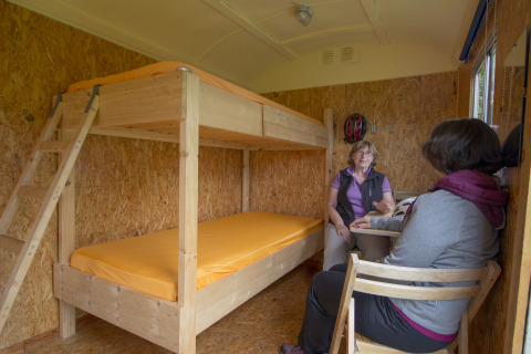 Twee vrouwen zitten te praten in een gezellige glamping pipowagen met houten stapelbedden en oranje matrassen.