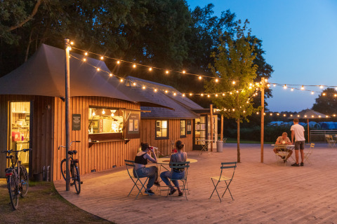 Outdoor dining area at Camping Huttopia De Roos glamping site with string lights and guests in the evening.