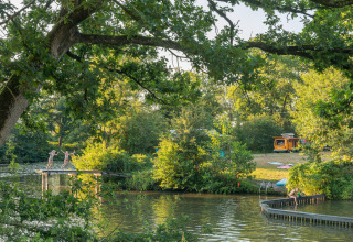Children play by the river at Camping Huttopia De Roos in Overijssel, camper van and woodland in the background.
