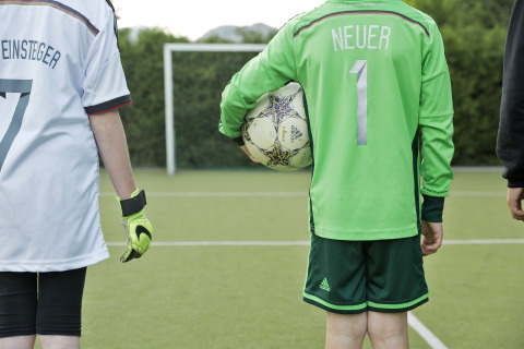 Zwei Kinder in deutschen Fußballtrikots auf dem Spielfeld, eines hält einen Ball, Sicht von hinten.
