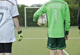 Zwei Kinder in deutschen Fußballtrikots auf dem Spielfeld, eines hält einen Ball, Sicht von hinten.