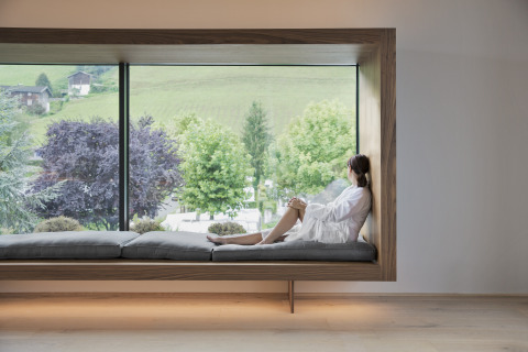 Woman in bathrobe relaxing at a large window seat with scenic views at Natürlich Hell Camping & Aparthotel, Tirol.