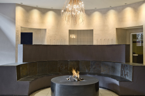 Lounge moderna con panchina circolare, camino centrale e lampadario presso Natürlich Hell Camping & Aparthotel.