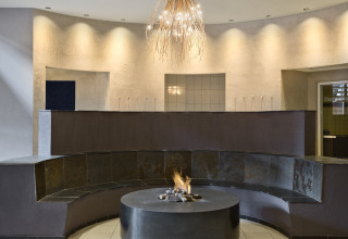 Lounge moderna con panchina circolare, camino centrale e lampadario presso Natürlich Hell Camping & Aparthotel.