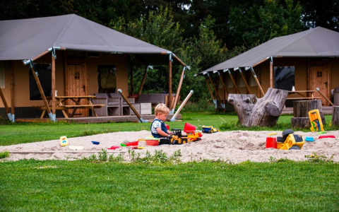 Un niño juega en una caja de arena con juguetes frente a las tiendas glamping en Glamping de Bosweide, Noord-Brabant.