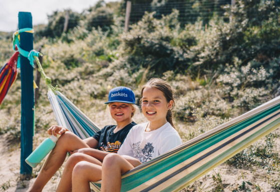 Dos niños sonrientes descansan en una hamaca en Surfana Family Camp Bloemendaal, Glamping Noord-Holland.