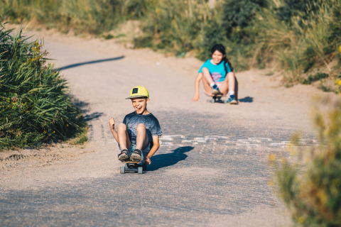 To børn kører på skateboards ned ad en grusvej ved Surfana Family Camp Bloemendaal, Glamping Noord-Holland.