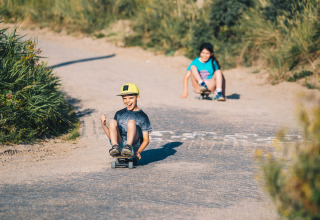Two kids riding skateboards on a sandy road at Surfana Family Camp Bloemendaal, Glamping Noord-Holland.