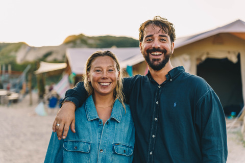 Deux adultes souriants sur la plage devant leur tente au Surfana Family Camp Bloemendaal Glamping Noord-Holland.