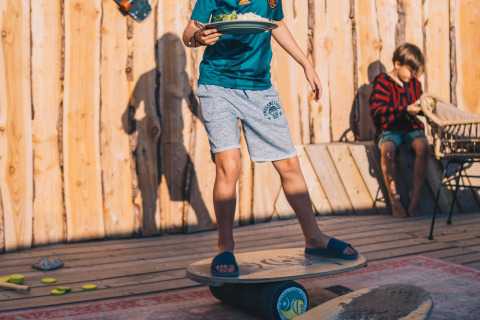 Ein Kind balanciert auf einem Balanceboard mit einem Teller Essen, während ein anderes im Hintergrund im Surfana Family Camp Bloemendaal sitzt.