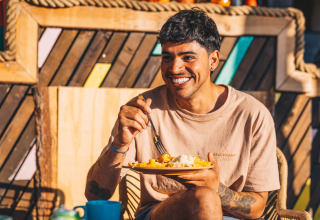 Giovane uomo sorride e mangia all'aperto al Surfana Family Camp Bloemendaal - Glamping Noord-Holland.