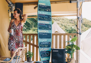 Femme tenant un café près d’un panneau d’événements à Surfana Family Camp Bloemendaal - Glamping Noord-Holland.