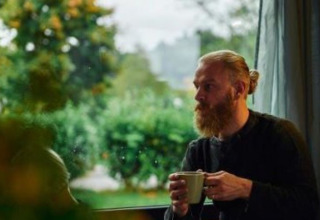 Bearded man sipping coffee in Wellnester Camping by Losheim am See, Tiny houses Saarland with a green view.