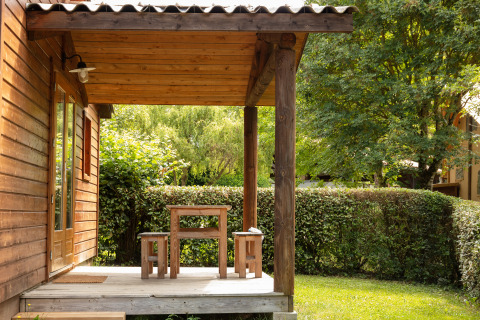 Cabaña de madera con porche y bancos en Camping Pré Fixe - Lodges en los Pirineos, rodeado de vegetación.