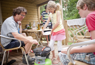 Famiglia che fa una grigliata insieme su una terrazza in legno a Huttopia Lac de Sillé - Glamping Pays de la Loire.