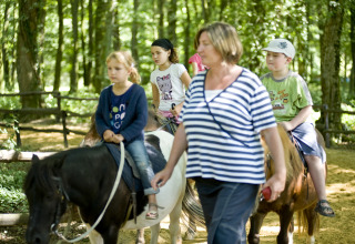 Børn rider på ponyer med en voksen leder i en skov, en aktivitet hos Huttopia Lac de Sillé glamping.
