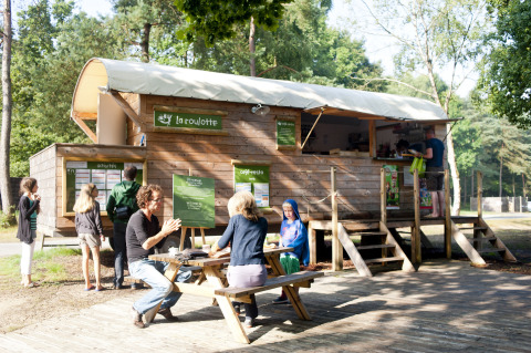 Glamping bij Huttopia Lac de Sillé, mensen aan picknicktafels en houten foodtruck in een bosrijke omgeving.