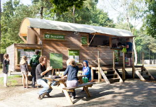 Glamping bij Huttopia Lac de Sillé, mensen aan picknicktafels en houten foodtruck in een bosrijke omgeving.